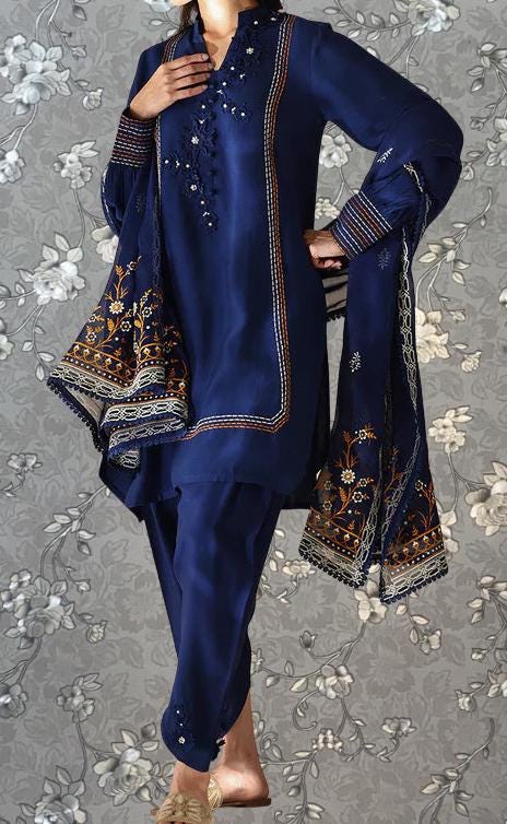 A person wearing a navy blue dress with intricate embroidery and gold accents, standing against a gray floral background.