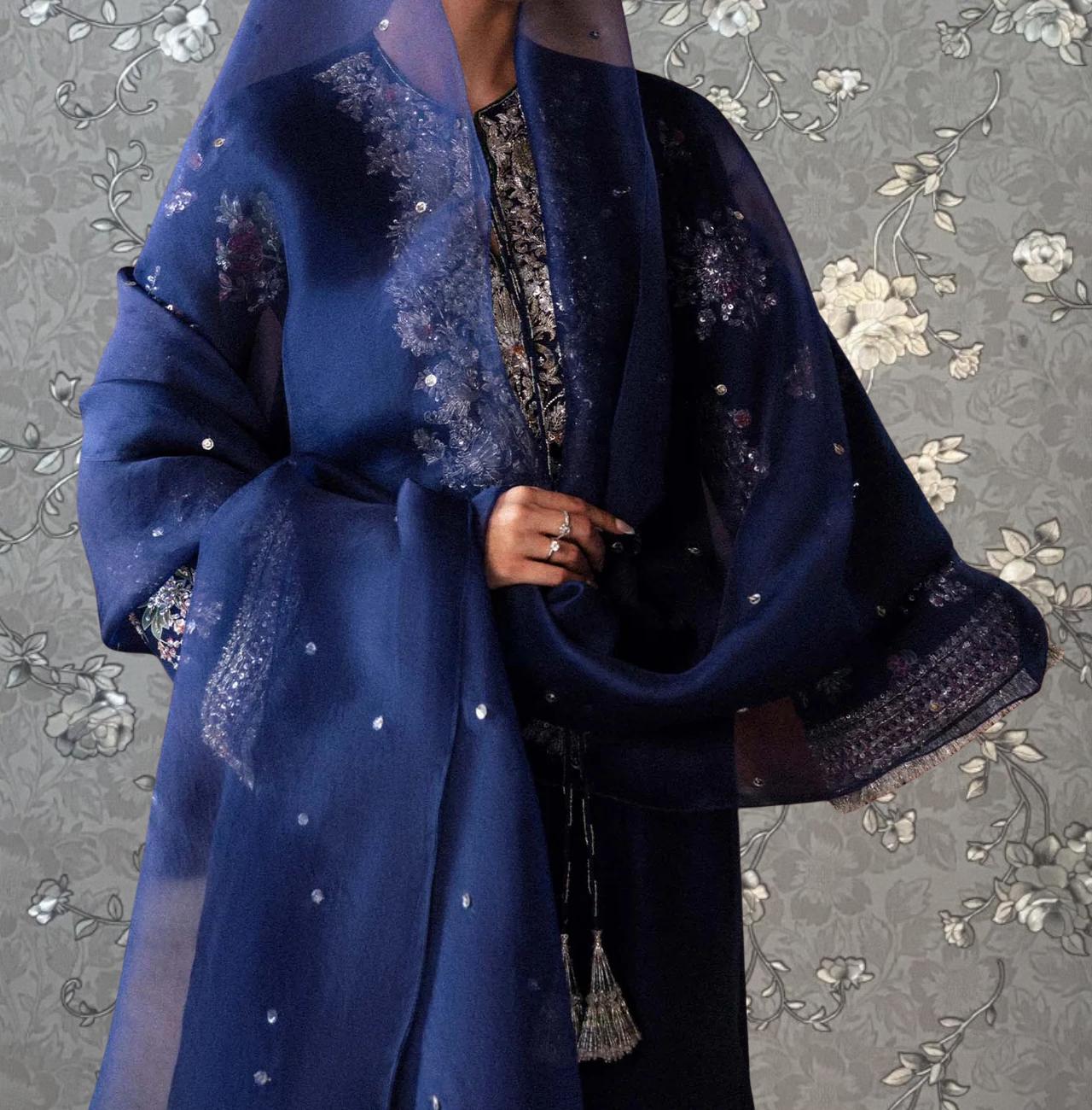 A person wearing a blue and white patterned dress with a matching blue shawl, standing against a gray floral background.