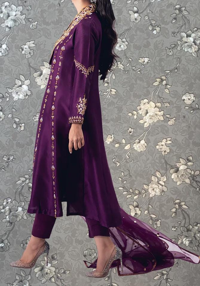 A woman wearing a long purple dress with gold embroidery and high heels is standing against a gray floral background.