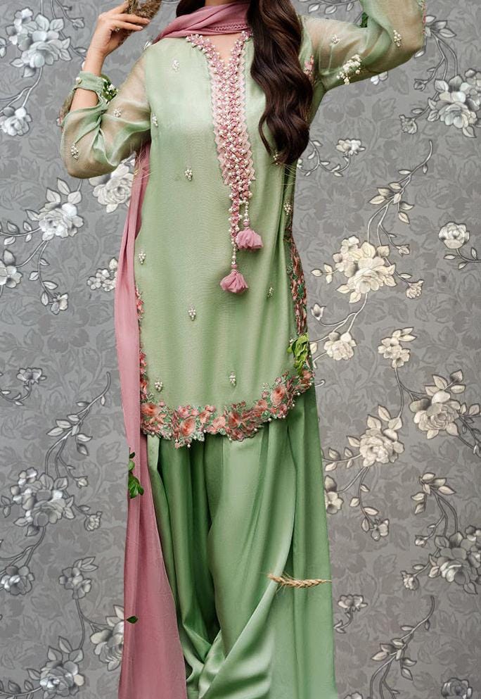 A woman wearing a green and pink dress with floral embroidery and a pink scarf, standing against a gray floral background.