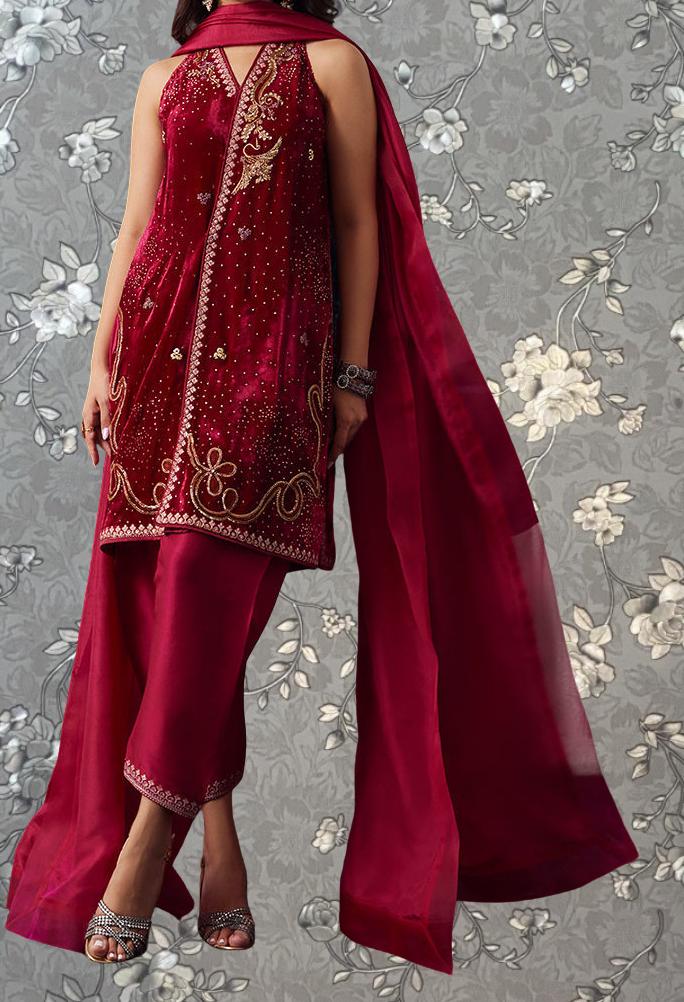 A woman wearing a red and gold embroidered dress with a matching red shawl, standing against a gray floral background.