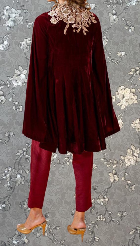 A person wearing a burgundy velvet cape with lace trim, paired with red pants, stands against a gray floral background.