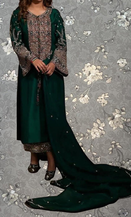 A woman wearing a green and gold embroidered dress with a long train, standing against a gray floral background.