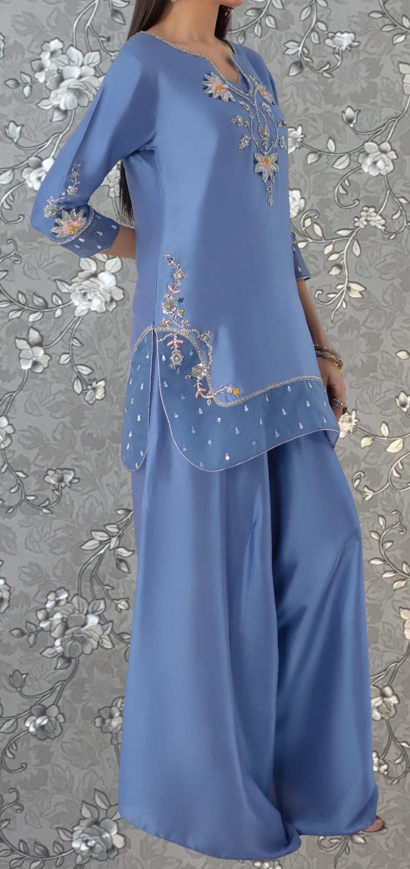 A woman wearing a blue dress with intricate embroidered designs on the front and sides, standing against a gray floral background.
