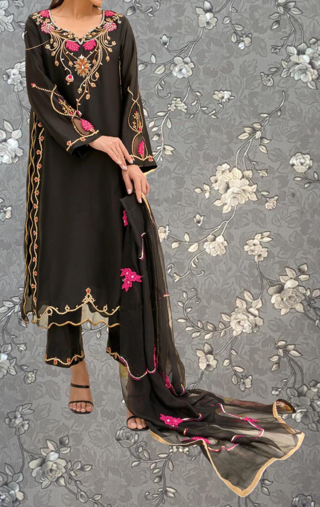 A woman wearing a black dress with gold embroidery and pink accents, standing against a gray floral background.