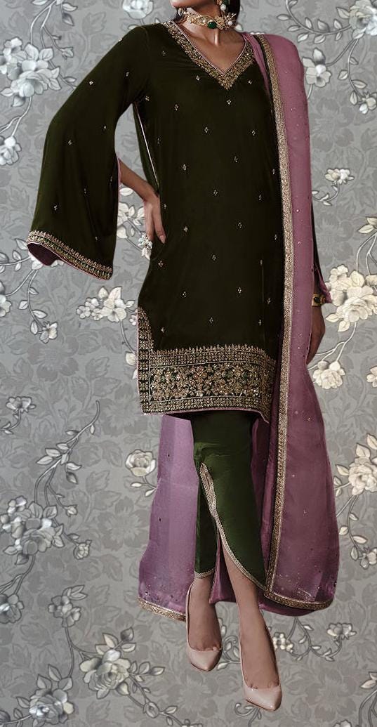 A woman wearing a dark green dress with gold embroidery and a pink shawl, standing against a gray floral background.