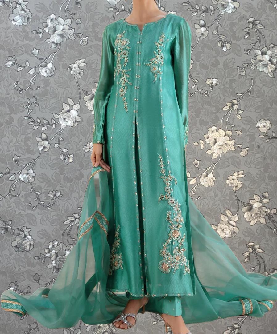A woman wearing a long, green dress with intricate embroidery stands against a gray floral background.