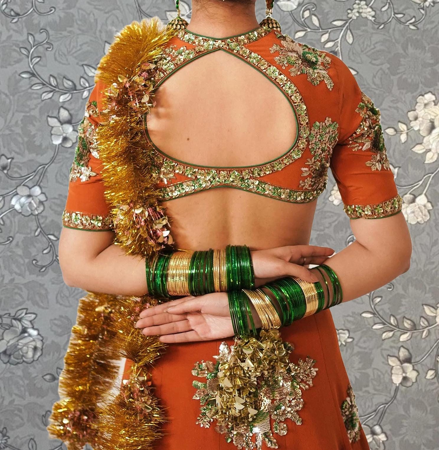 A woman wearing a vibrant orange and green dress with a large gold and green fringe draped over her back. She is also wearing multiple bracelets and earrings.