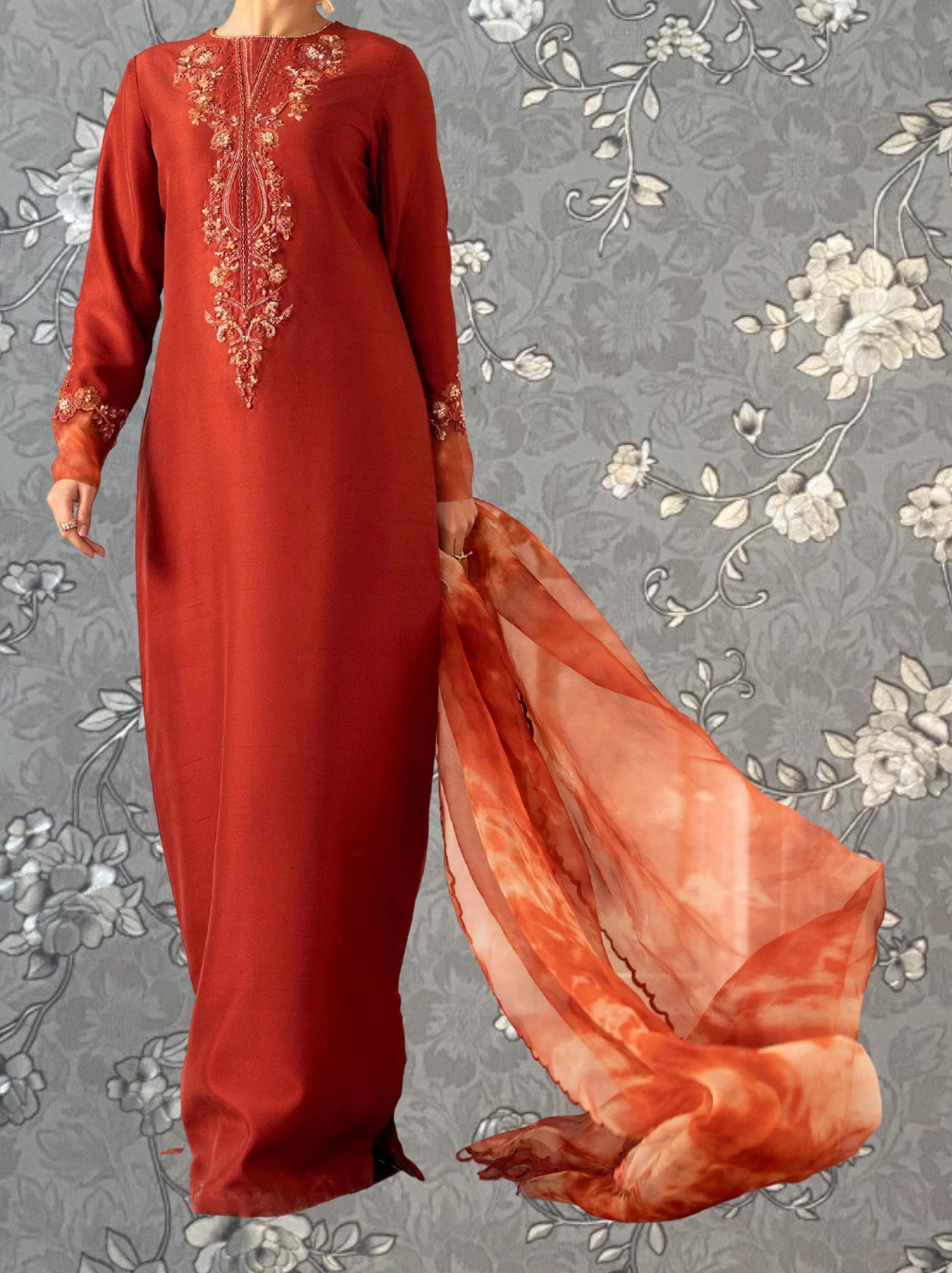 A woman wearing a long, red dress with intricate embroidery stands against a gray floral background.