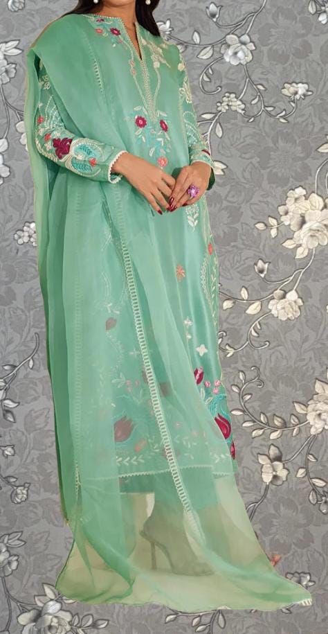 A woman wearing a long, green dress with floral embroidery stands against a gray floral background.