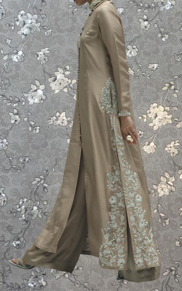 A person wearing a long, beige dress with intricate embroidered patterns stands against a gray floral background.