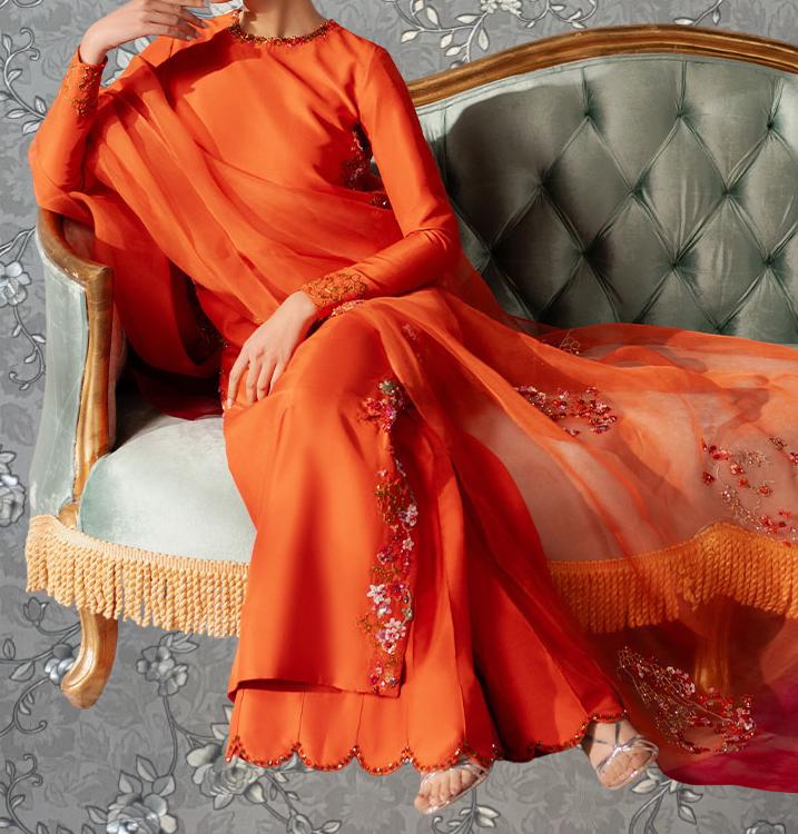 A person wearing an orange dress with floral embroidery is sitting on a green upholstered chair with gold fringe.