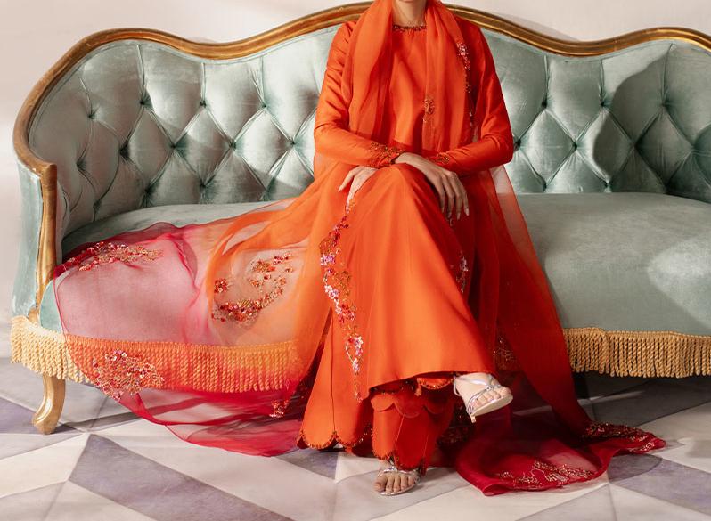 A person wearing an orange dress with a long train and a matching red scarf is sitting on a green velvet couch with gold trim.
