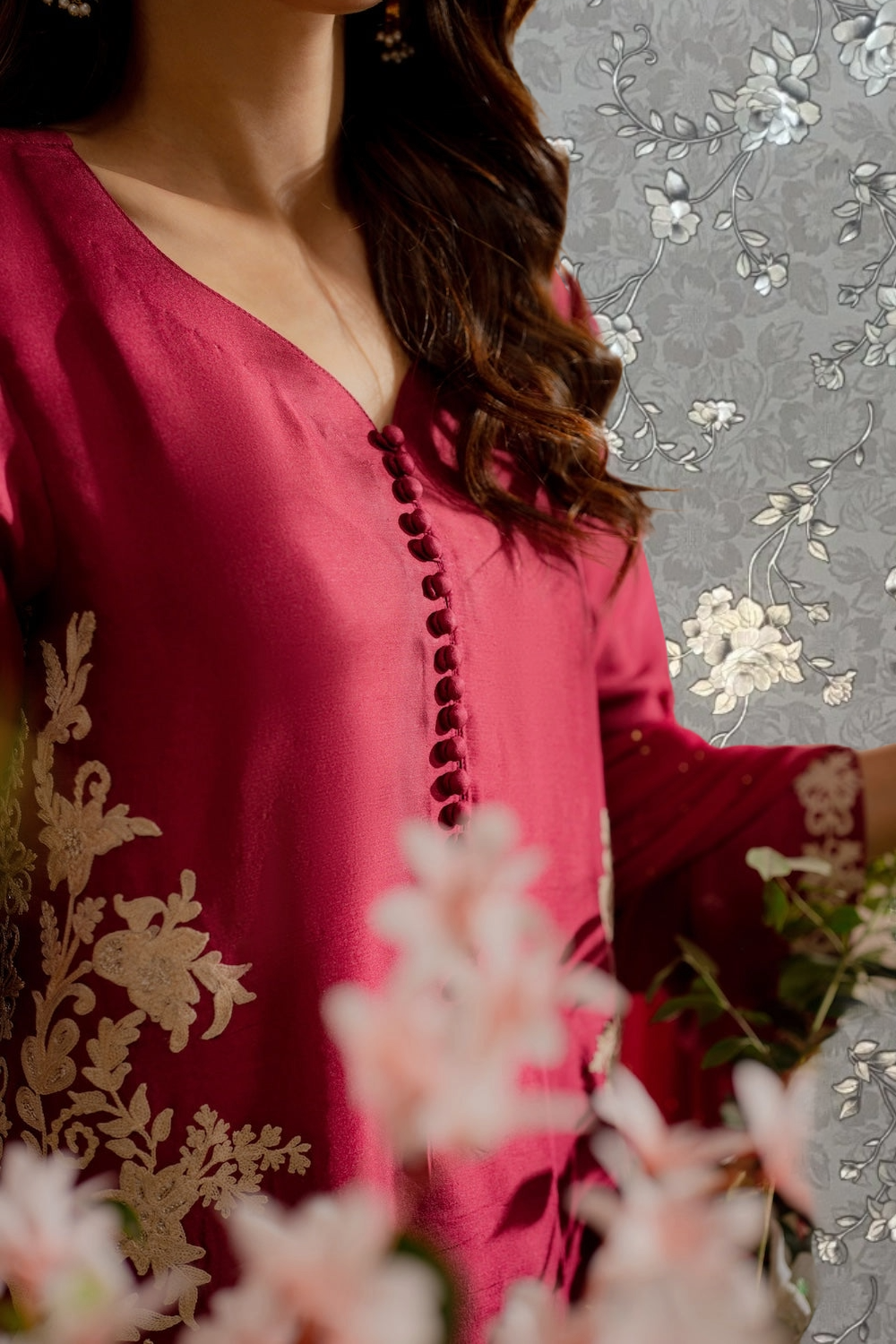 A close-up of a woman wearing a red dress with floral embroidery, standing in front of a floral patterned background.