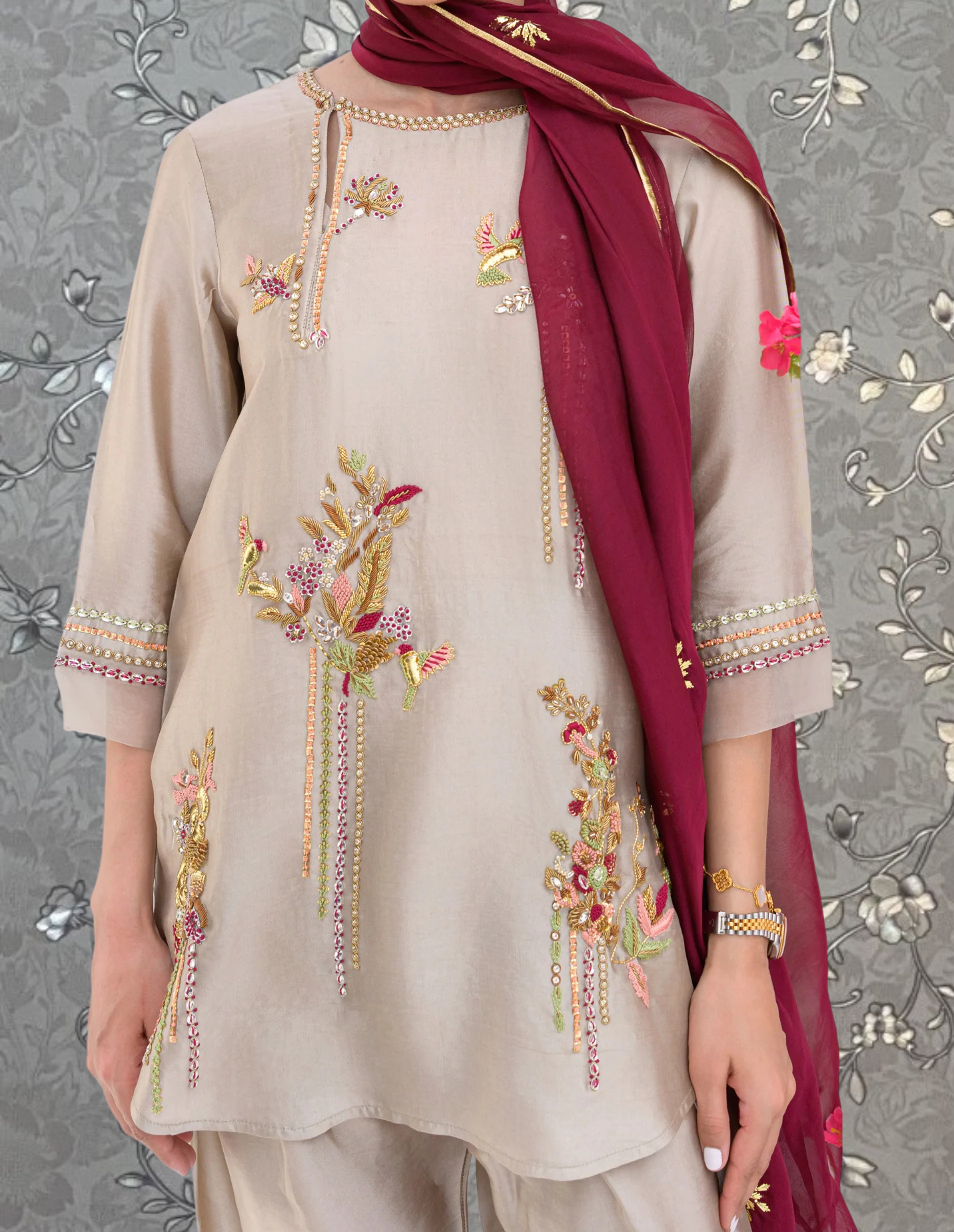 A person wearing a beige dress with intricate embroidered designs, including flowers and leaves, and a red scarf draped over their shoulders.
