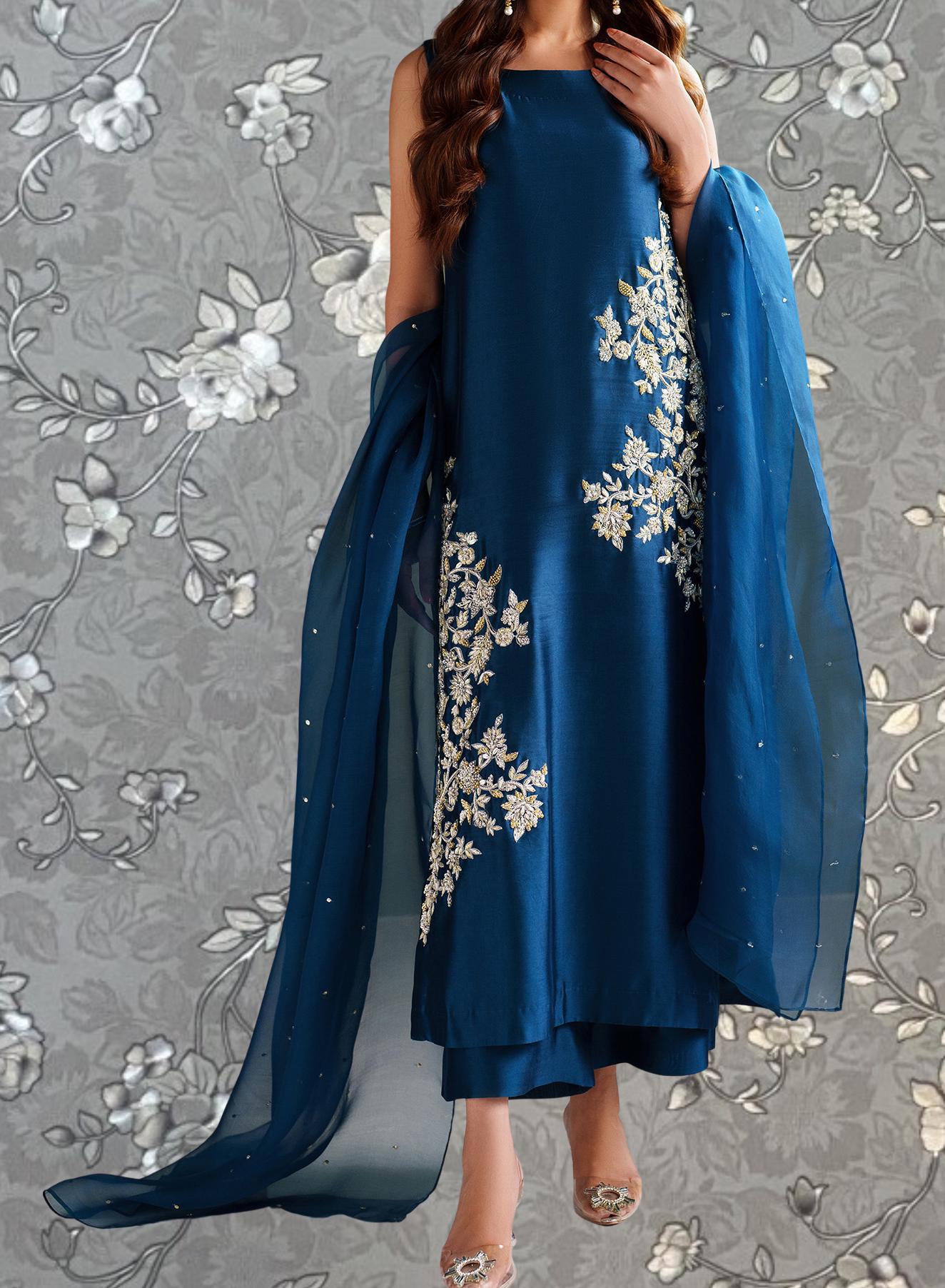 A woman wearing a blue dress with white floral embroidery stands against a gray floral background.