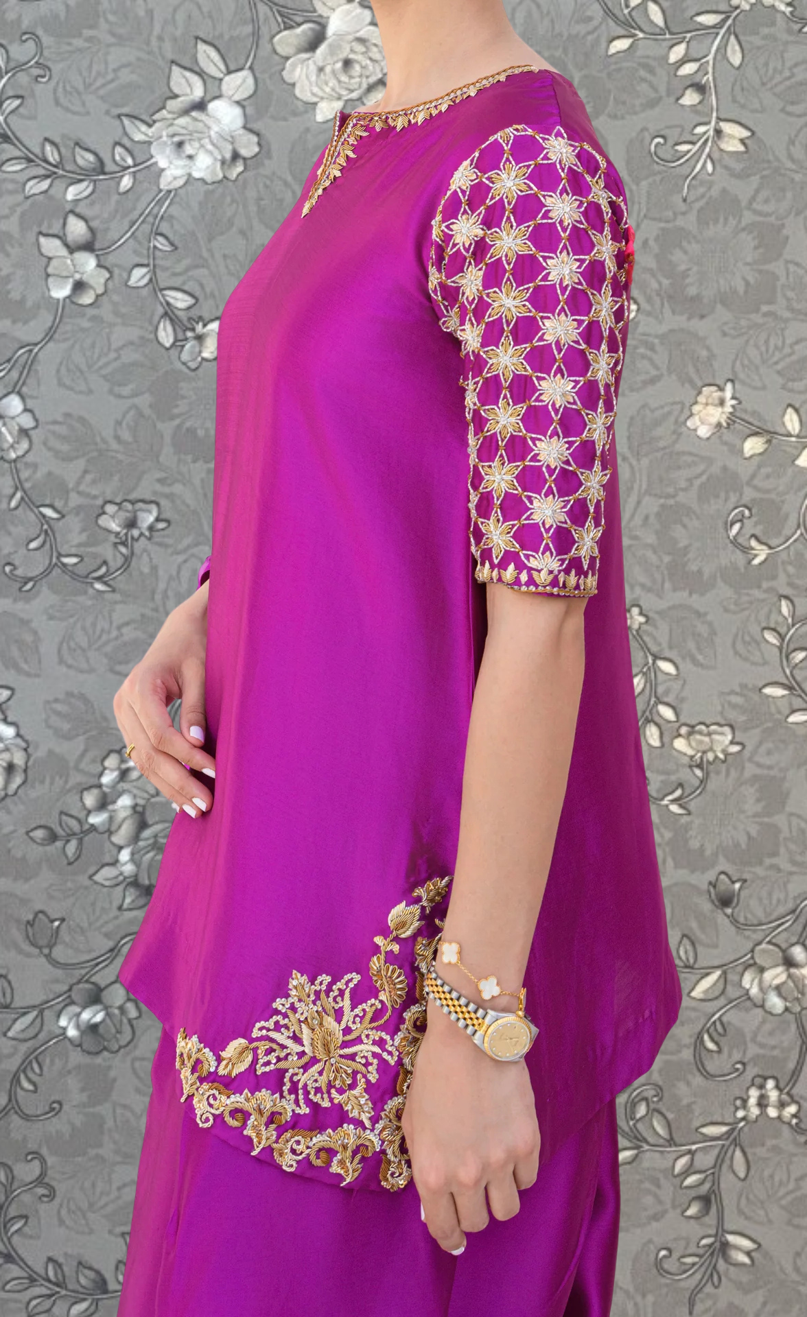 A woman wearing a vibrant purple dress with gold embroidery stands against a gray floral background.