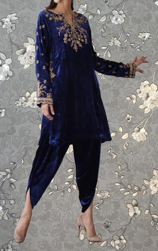 A woman wearing a blue velvet dress with gold embroidery and matching heels poses against a gray floral background.