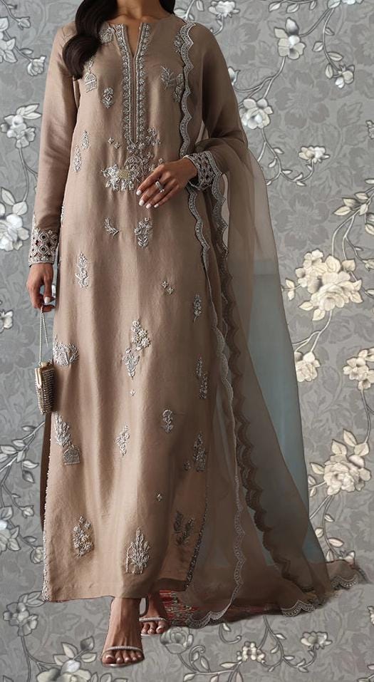 A woman wearing a long, beige dress with intricate embroidery stands against a gray floral background.