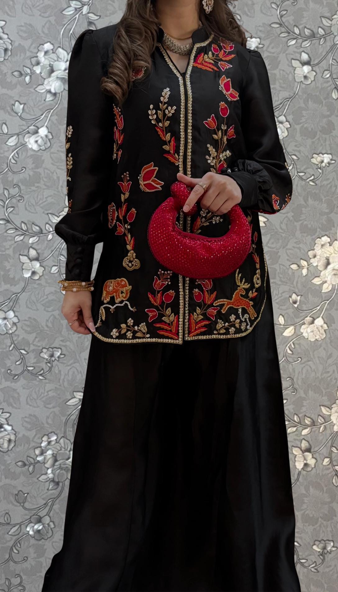A woman wearing a black dress with intricate embroidery and holding a red purse stands against a gray floral background.