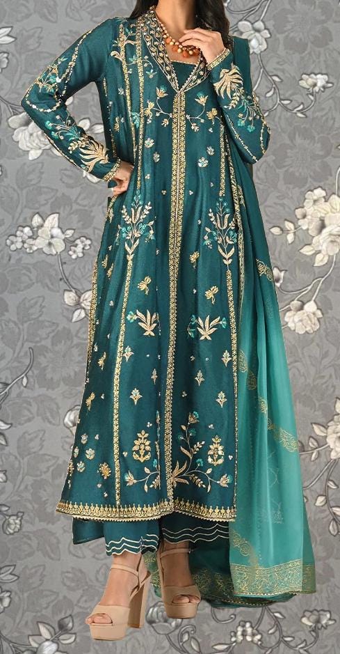 A woman wearing a long, green dress with gold embroidery and floral patterns, standing against a gray floral background.