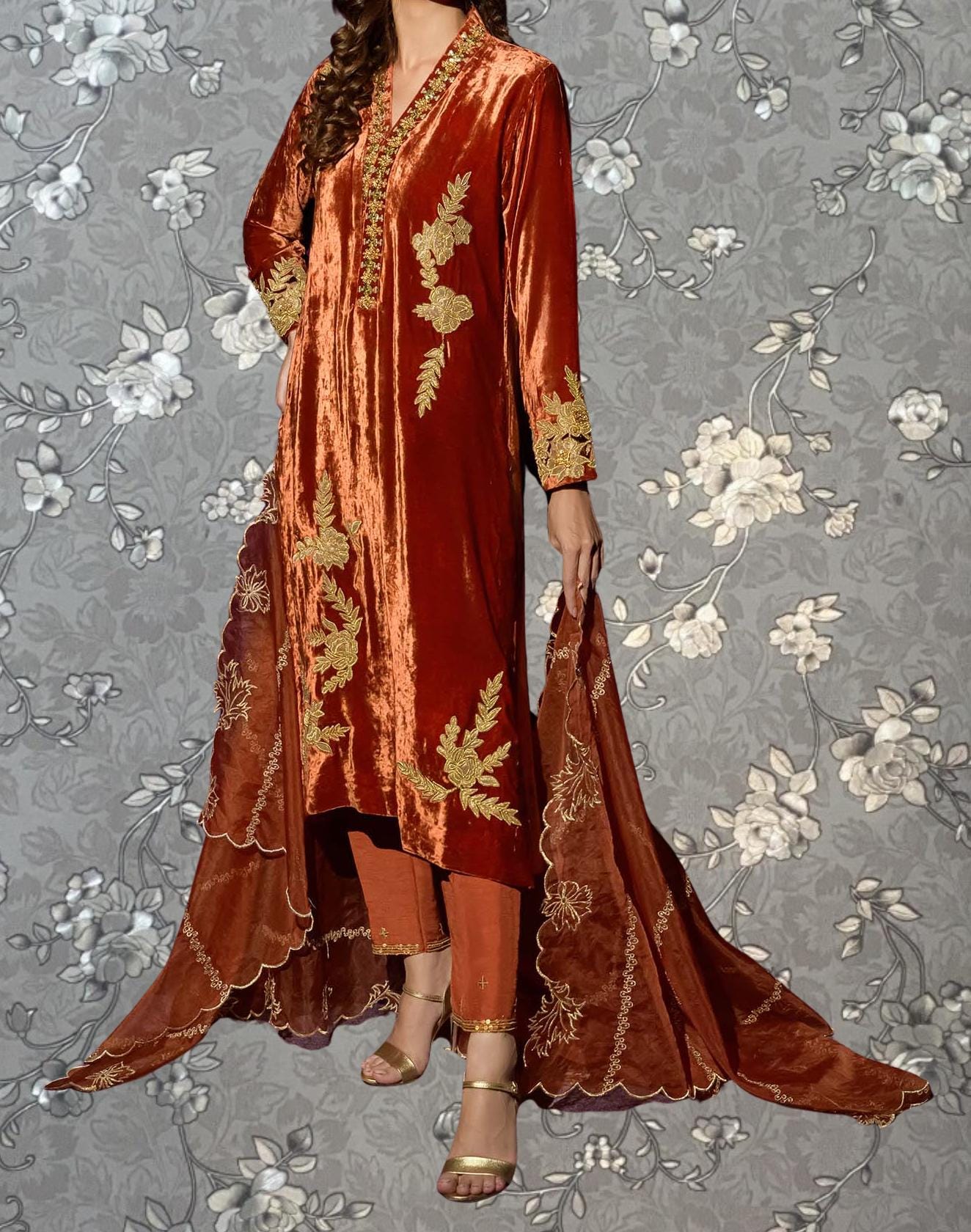 A woman wearing a long, red velvet dress with gold embroidery stands against a gray floral background.
