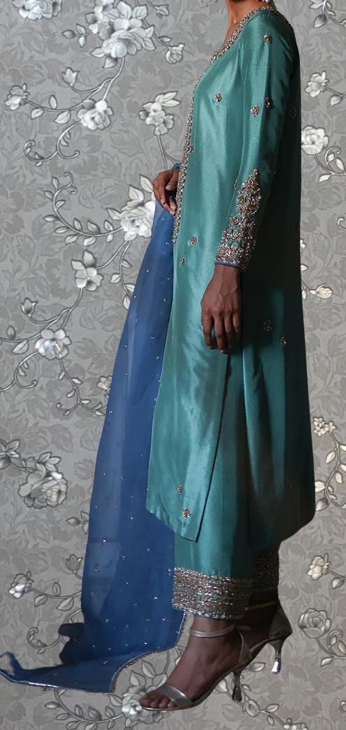 A person wearing a long, green dress with intricate embroidery and a matching blue shawl stands against a gray floral background.
