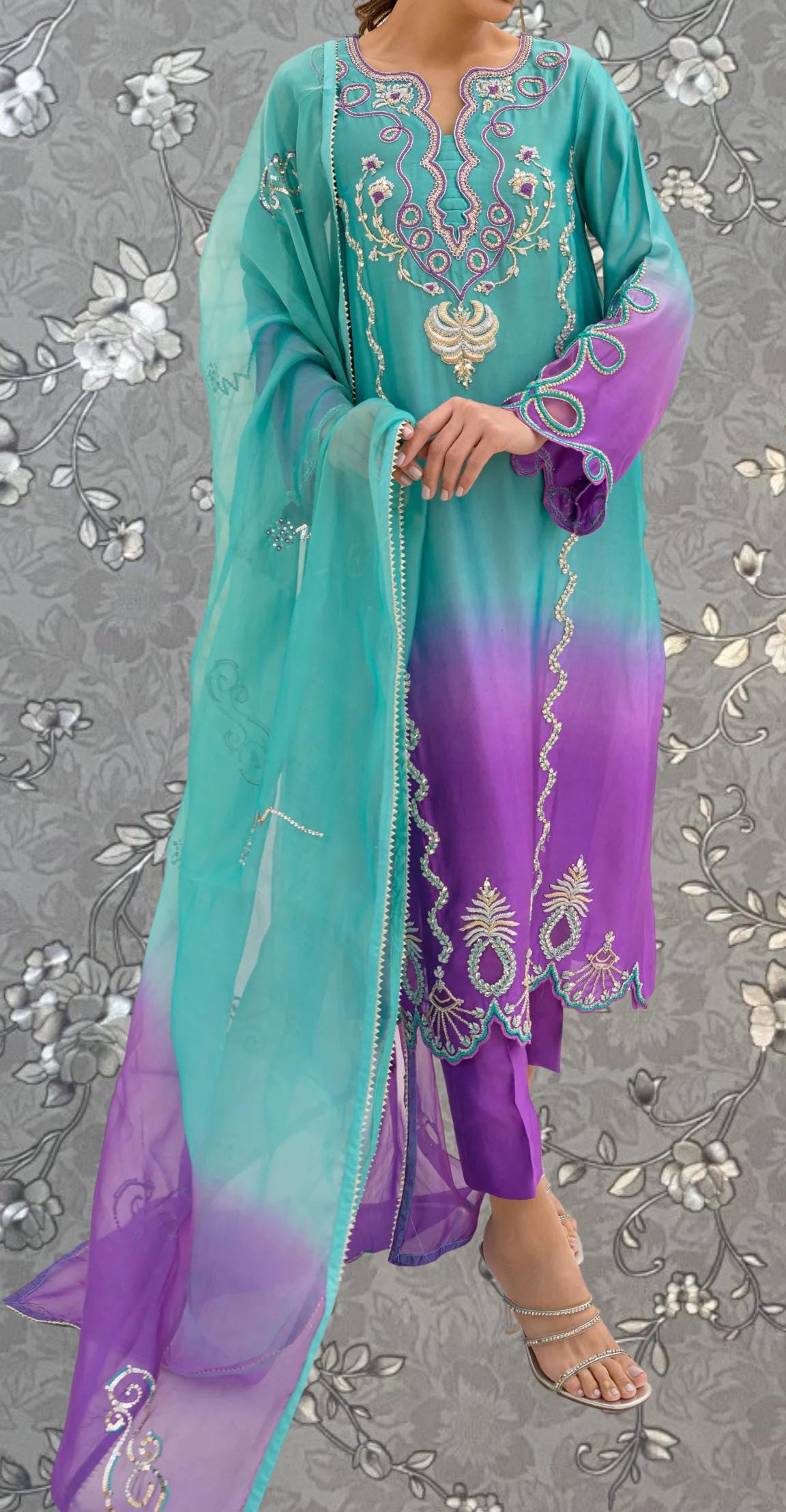 A woman wearing a colorful, patterned dress with a turquoise and purple color scheme, standing against a gray floral background.