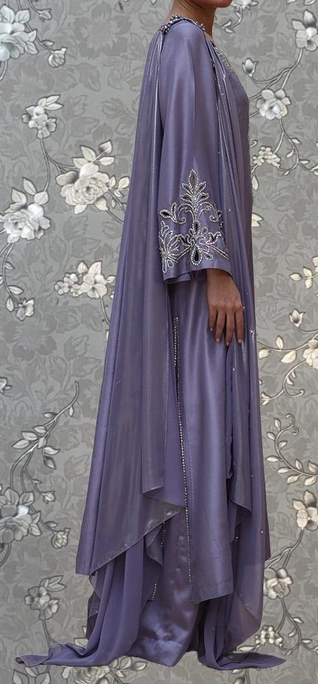 A person wearing a long, flowing purple dress with intricate embroidery stands against a gray floral background.