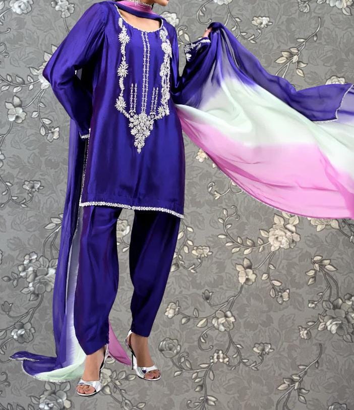 A person wearing a purple and pink patterned dress with a long, flowing skirt and high heels, standing against a gray floral background.
