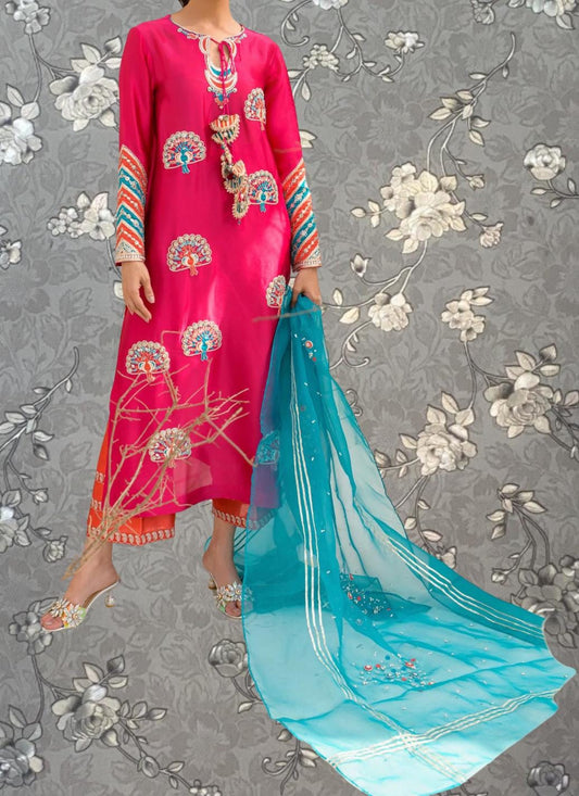 A woman wearing a bright pink dress with intricate floral embroidery and a matching turquoise shawl stands against a gray floral background.