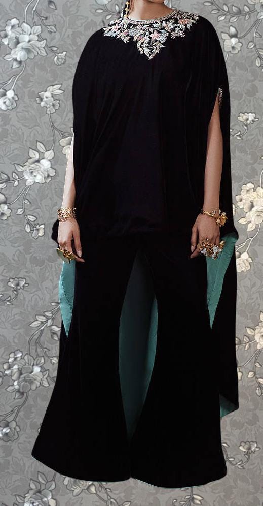 A person wearing a black cape-like garment with a floral pattern and gold accessories, standing against a gray floral background.