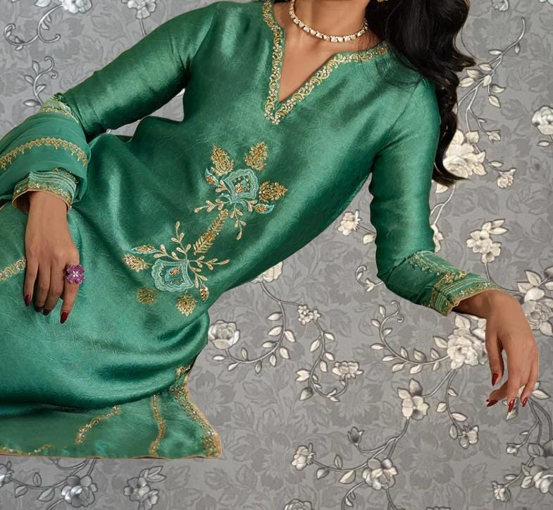 A woman wearing a green dress with gold embroidery and a necklace is shown against a gray floral background.