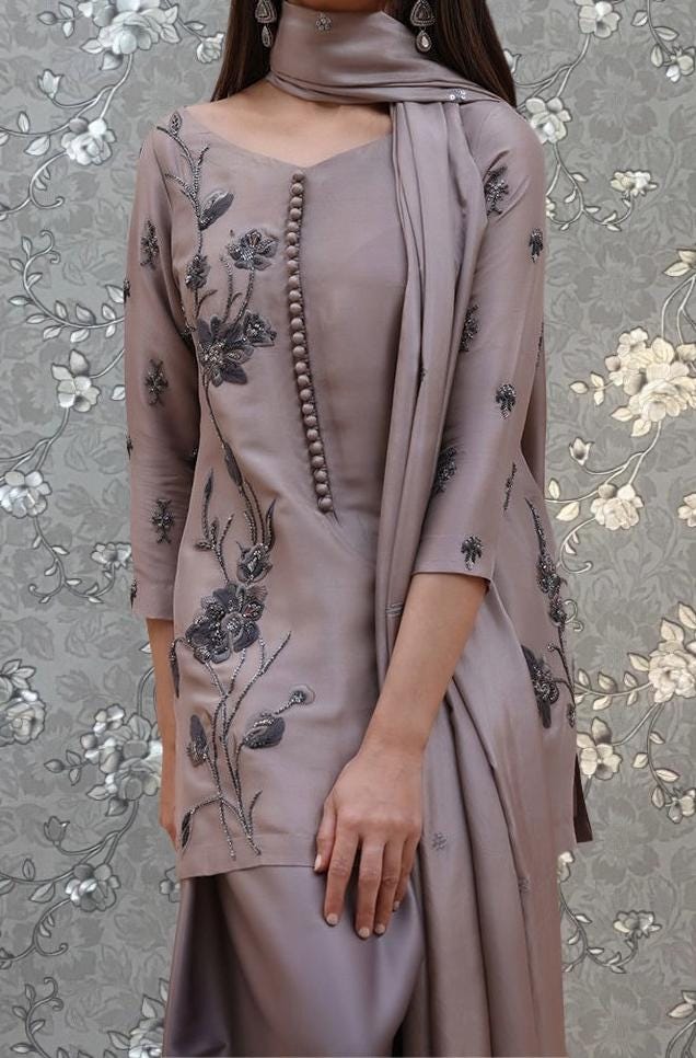 A woman wearing a gray dress with floral embroidery stands against a gray floral background.