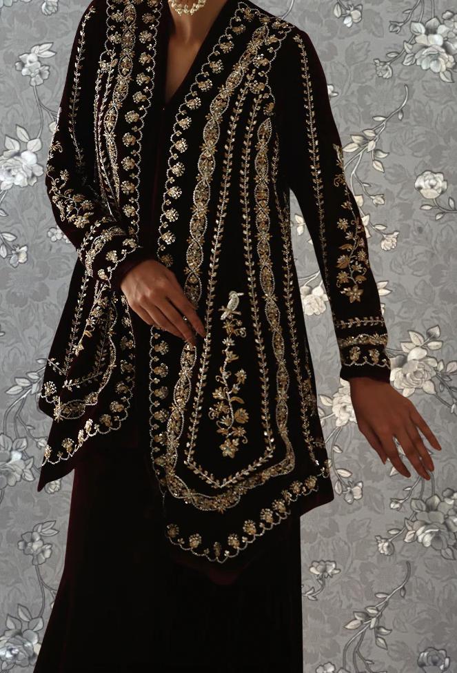 A person wearing an ornate, black and gold embroidered jacket stands against a floral patterned background.