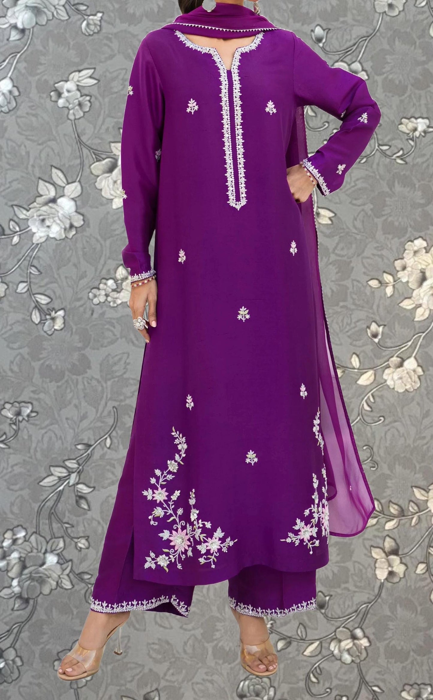 A woman wearing a long purple dress with white floral embroidery stands against a gray floral background.
