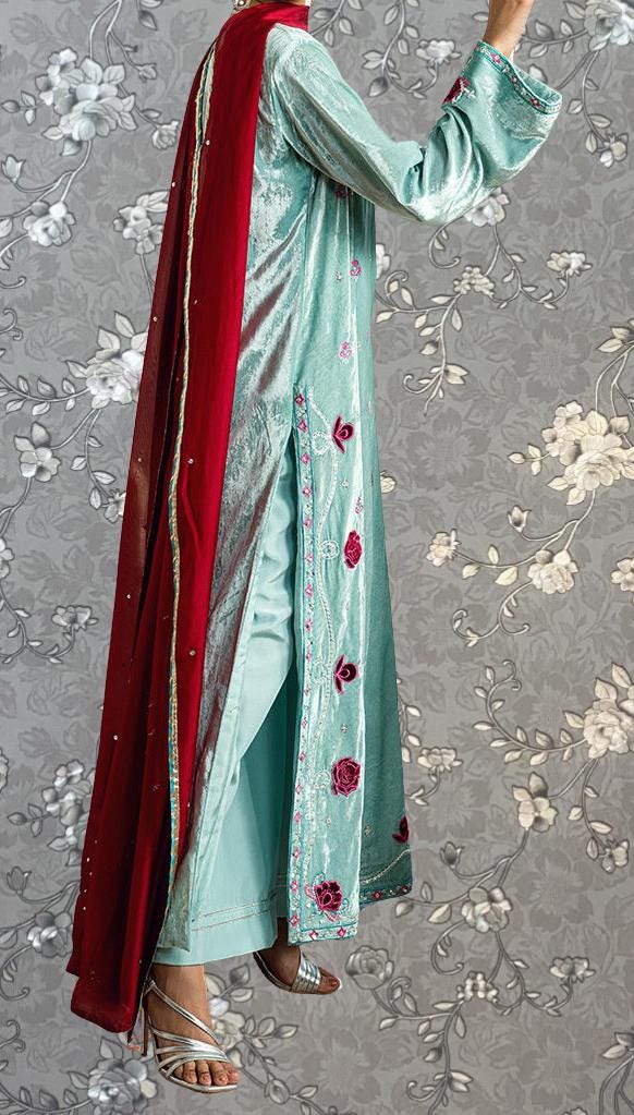 A person wearing a long, light blue dress with red accents and a matching red shawl, standing against a gray floral background.