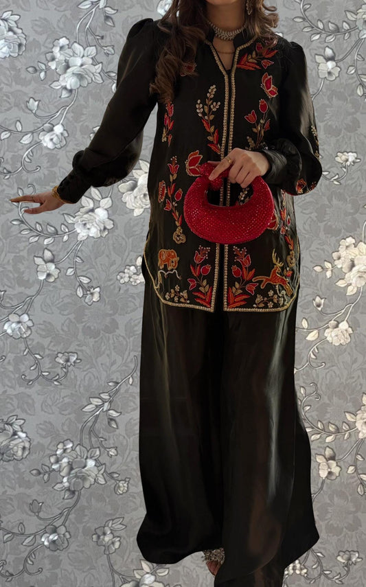 A woman wearing a black dress with red floral embroidery and holding a red purse stands against a gray floral background.