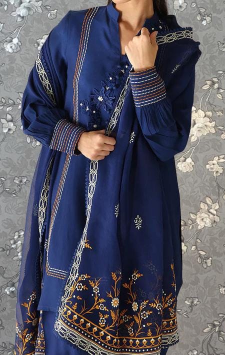 A woman wearing a blue dress with intricate embroidered patterns and designs, posing against a gray floral background.
