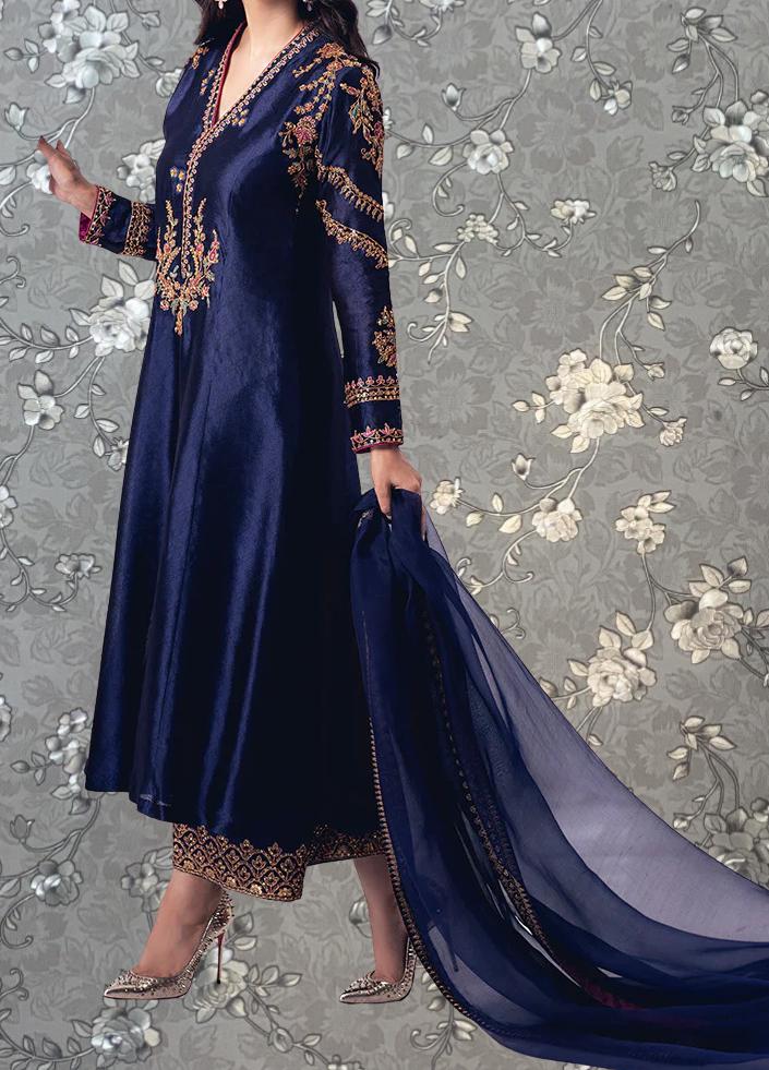 A woman wearing a long, navy blue dress with gold embroidery and a matching blue shawl, standing against a gray floral background.