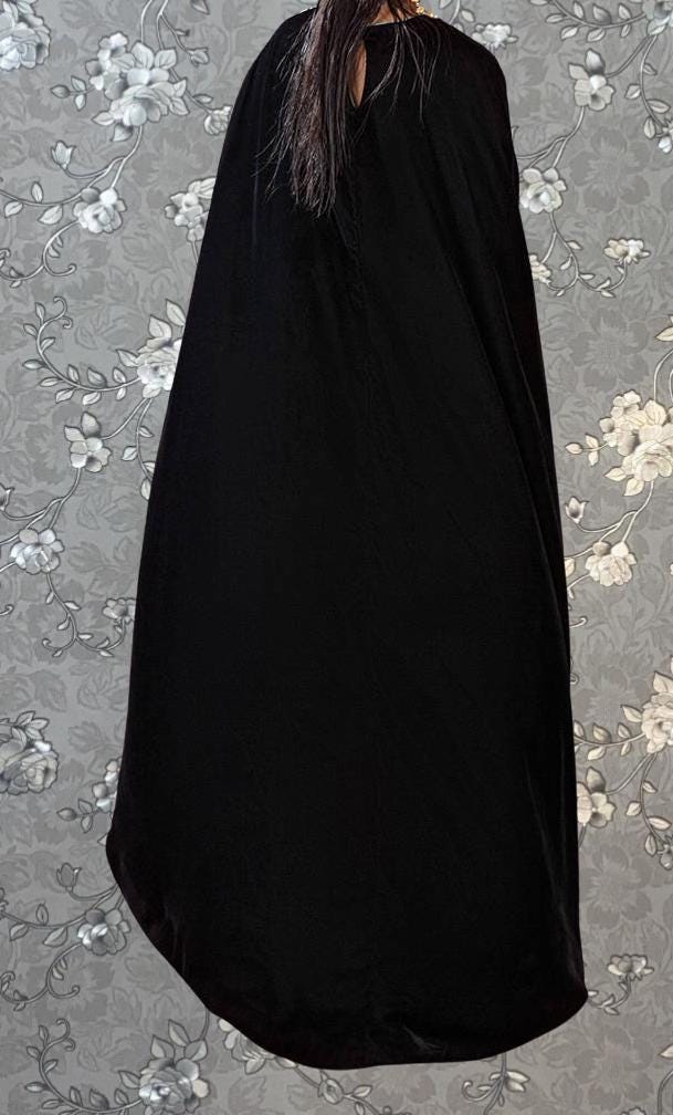 A person wearing a long black cape stands against a floral patterned background.