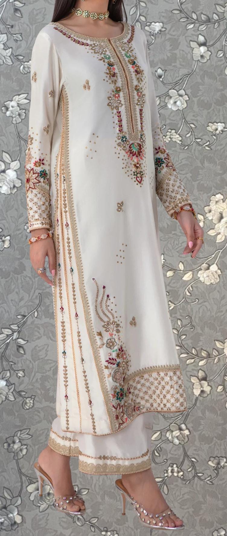 A woman wearing a white dress with intricate embroidery and gold accents, standing against a gray floral background.