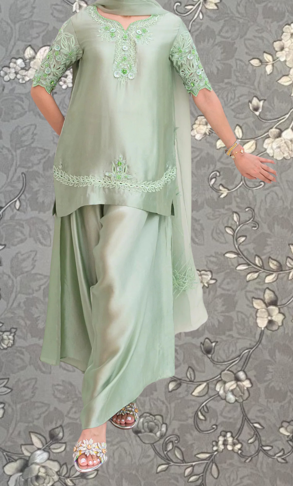 A woman wearing a green dress with intricate embroidery and floral patterns, standing against a gray floral background.