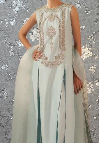 A woman wearing a light blue dress with intricate embroidery and a long train, posing against a gray floral background.