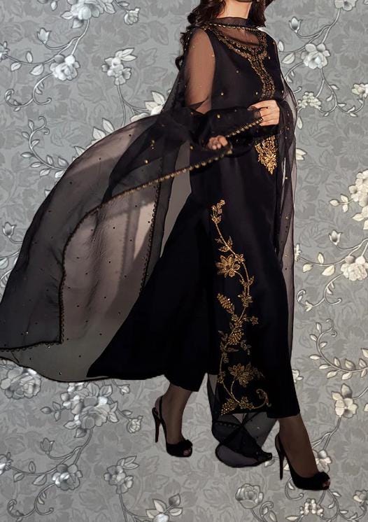 A woman wearing a long black dress with gold embroidery and a sheer black cape is standing against a floral background.