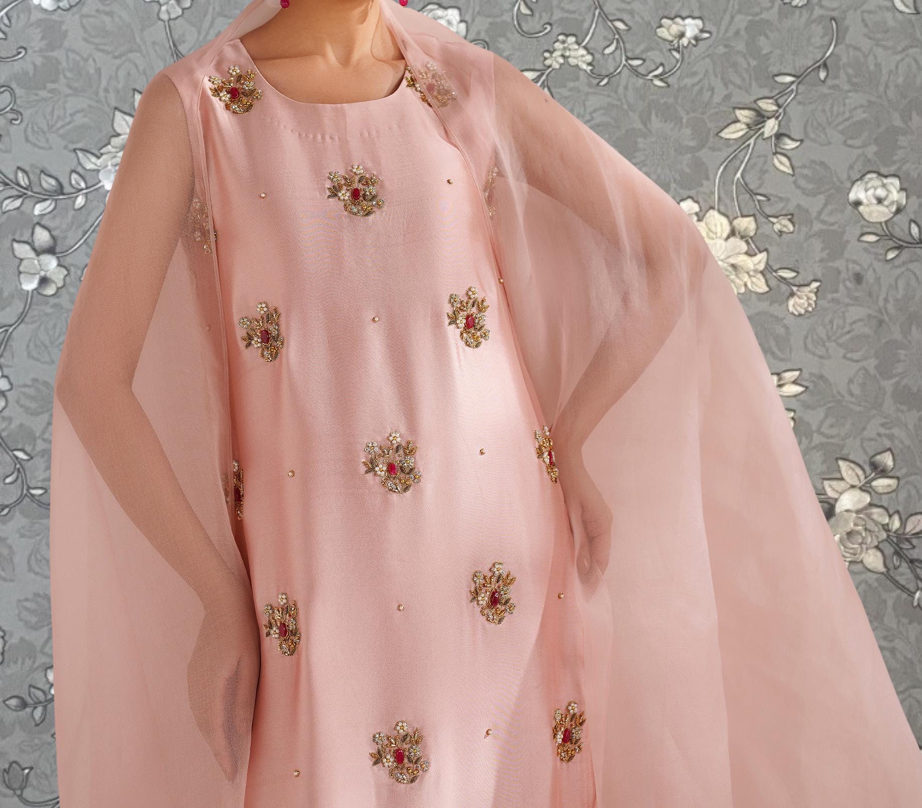 A close-up of a woman wearing a pink dress with gold embroidered flowers on it, standing against a gray floral background.