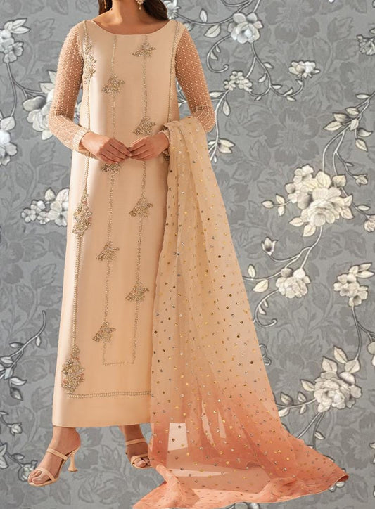 A woman wearing a long, beige dress with intricate embroidery and a matching shawl stands against a gray floral background.