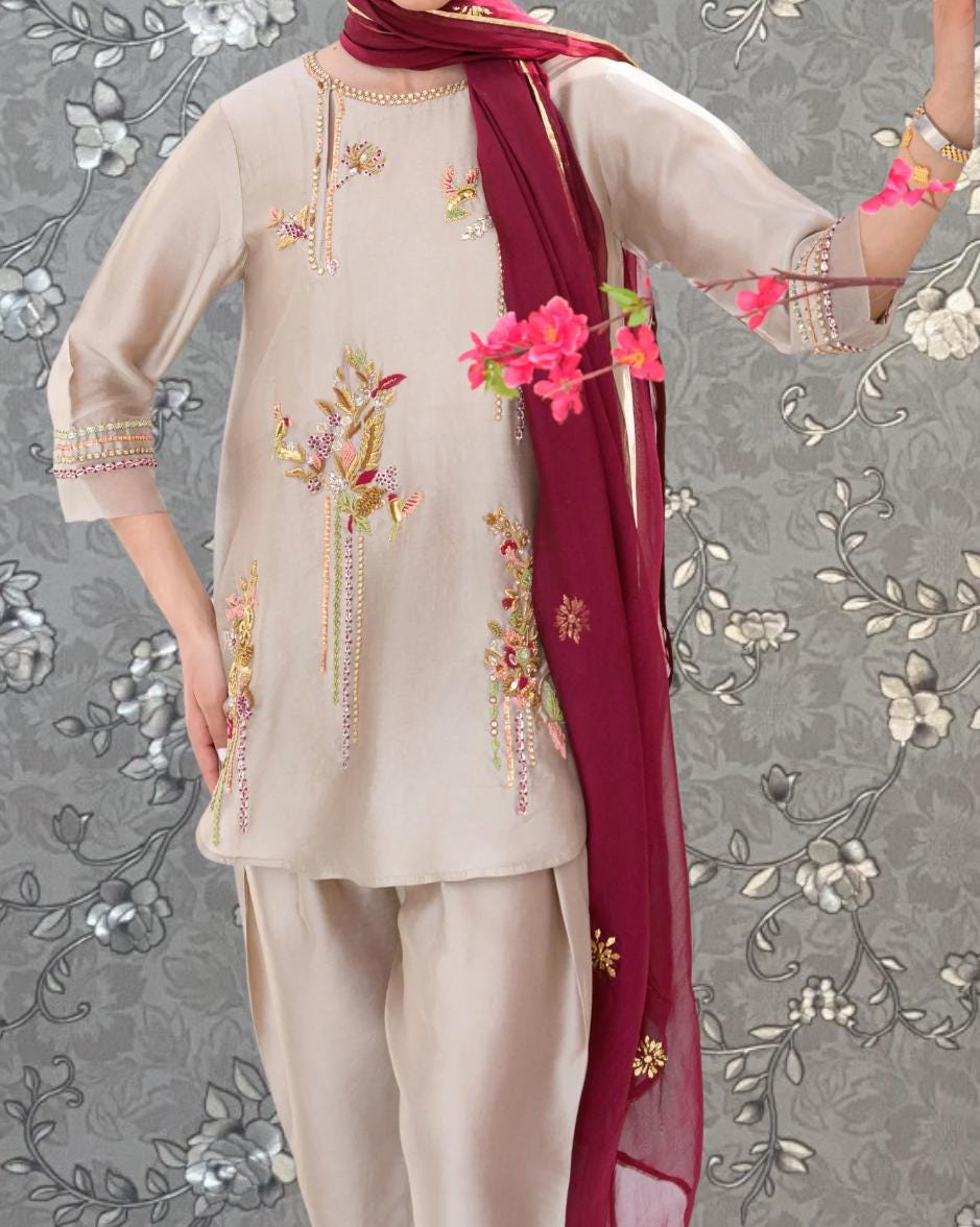 A person wearing a beige dress with floral embroidery and a red scarf, posing against a gray floral background.