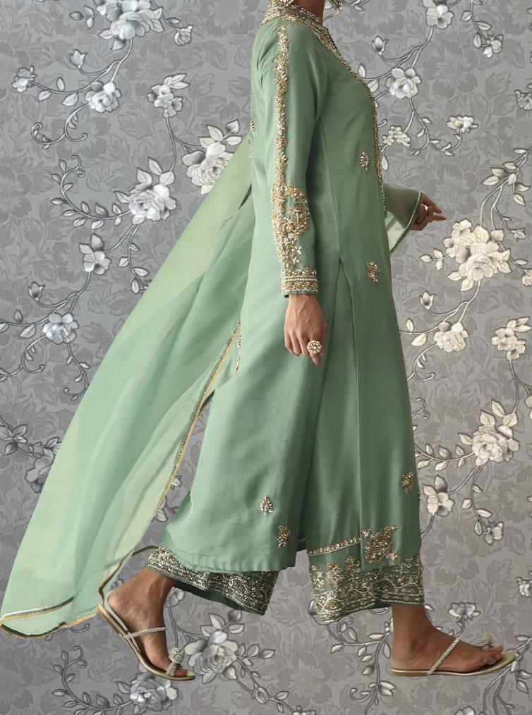 A woman wearing a long, green dress with gold embroidery and a matching green shawl is walking against a gray floral background.
