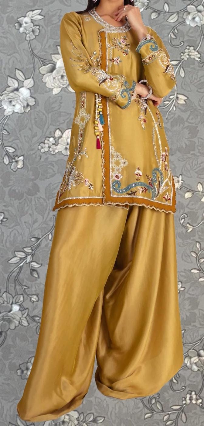 A woman wearing a gold and white patterned dress with intricate embroidery, standing against a gray floral background.